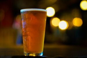 A pint glass of golden IPA with a thin white head on a bar, warm lights blurred behind it