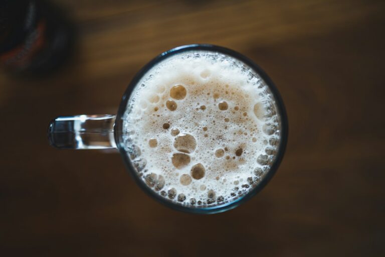 A glass mug of beer with a thick tan head, seen from above on a wooden table