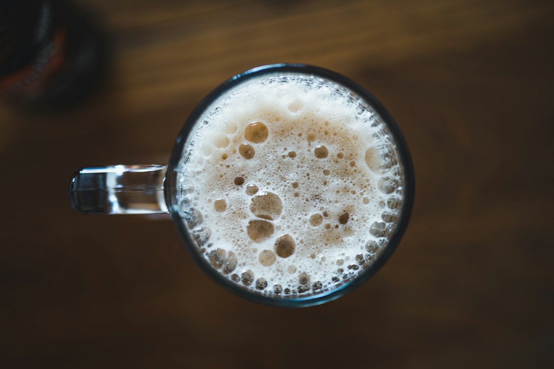 A glass mug of beer with a thick tan head, seen from above on a wooden table