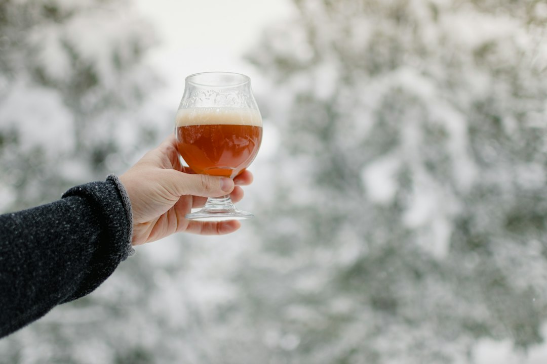 Hand holding a tulip glass of amber winter ale in front of snow-covered trees